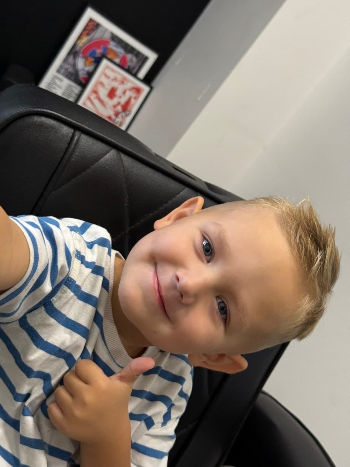 Young blonde boy in a blue striped shirt smiling and giving a thumbs up gesture.