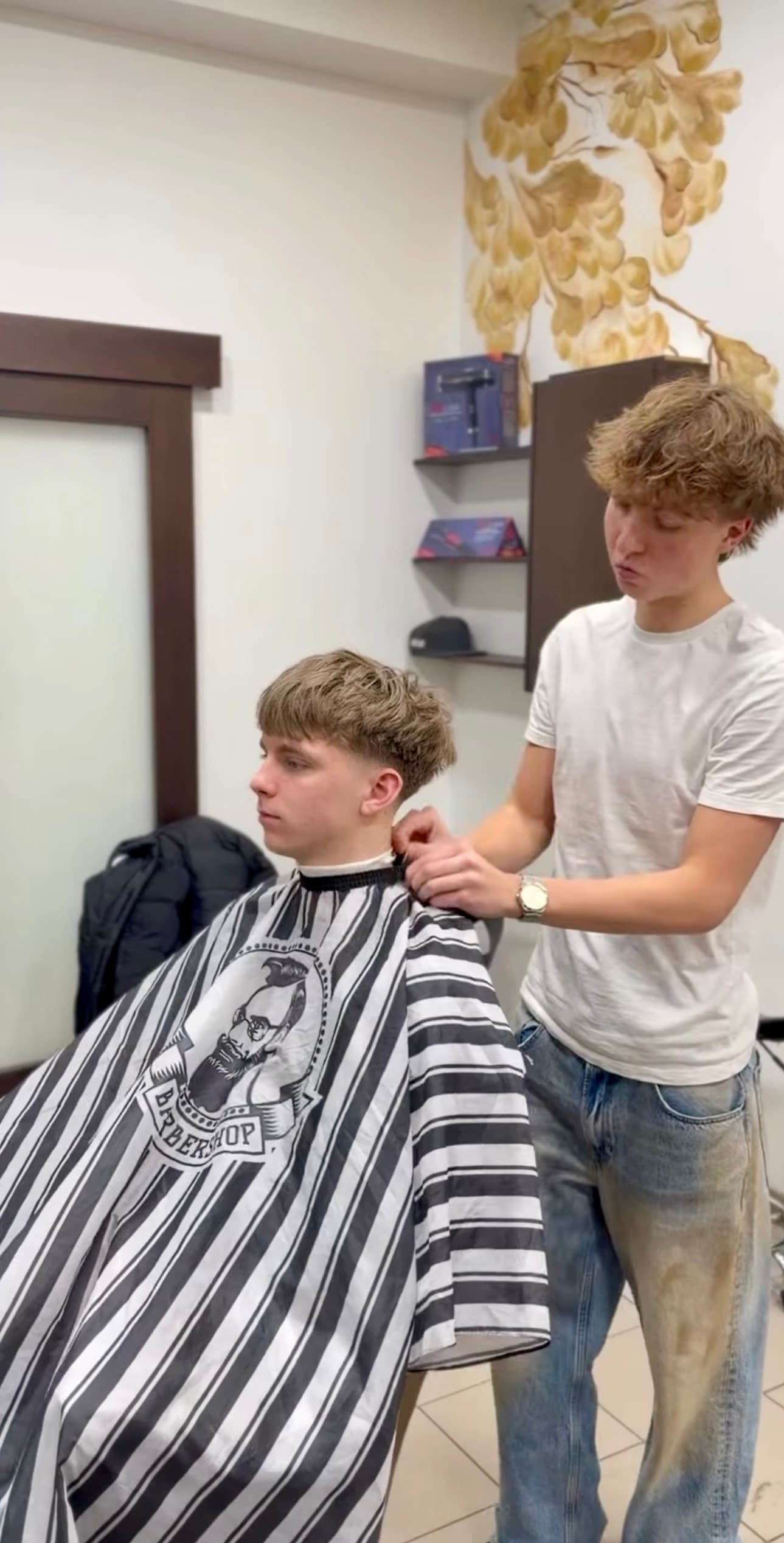 Young barber in a white t-shirt adjusts a striped cape on a seated client.