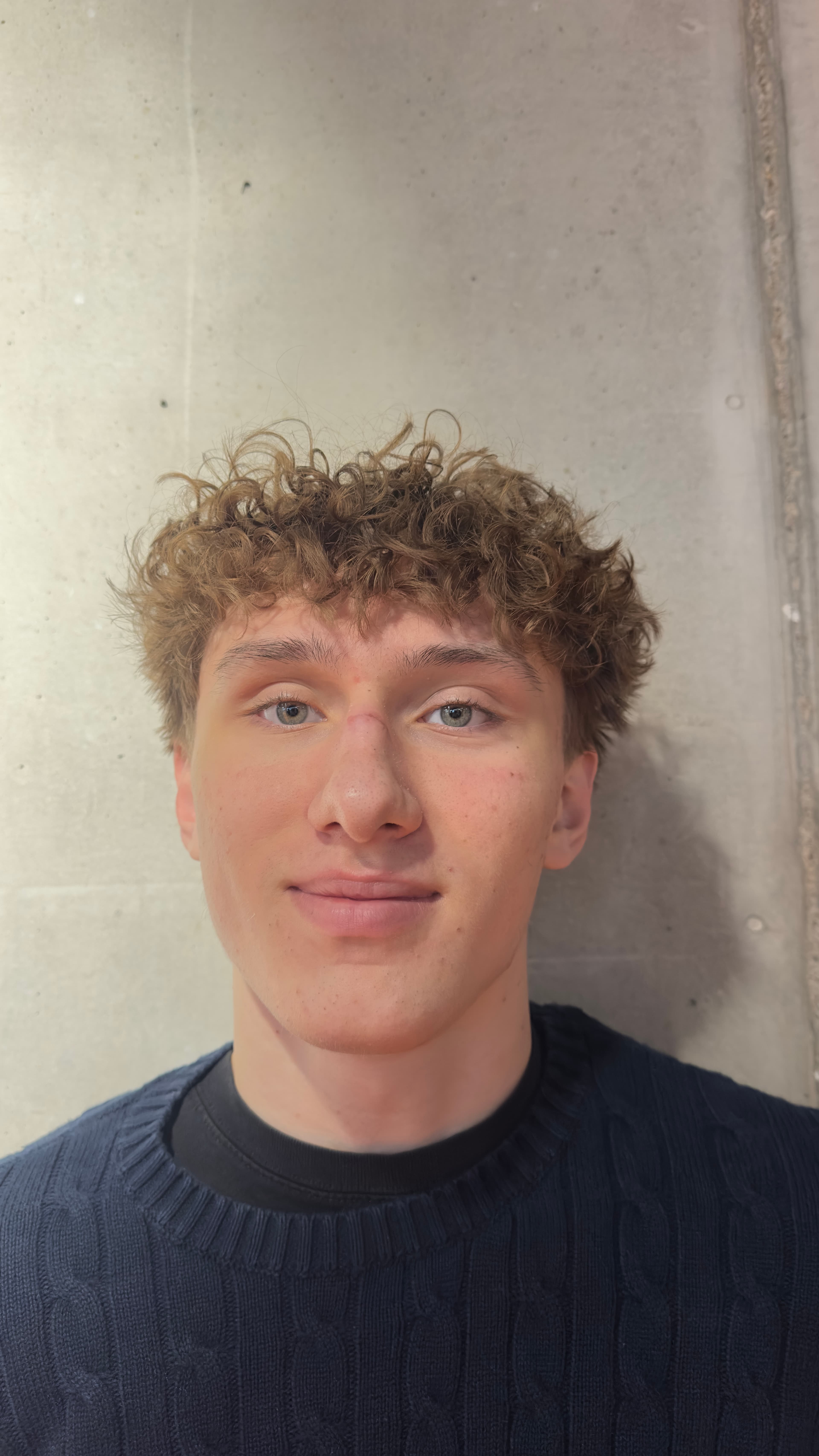 Young man with curly brown hair and blue eyes smiling against a plain concrete wall.
