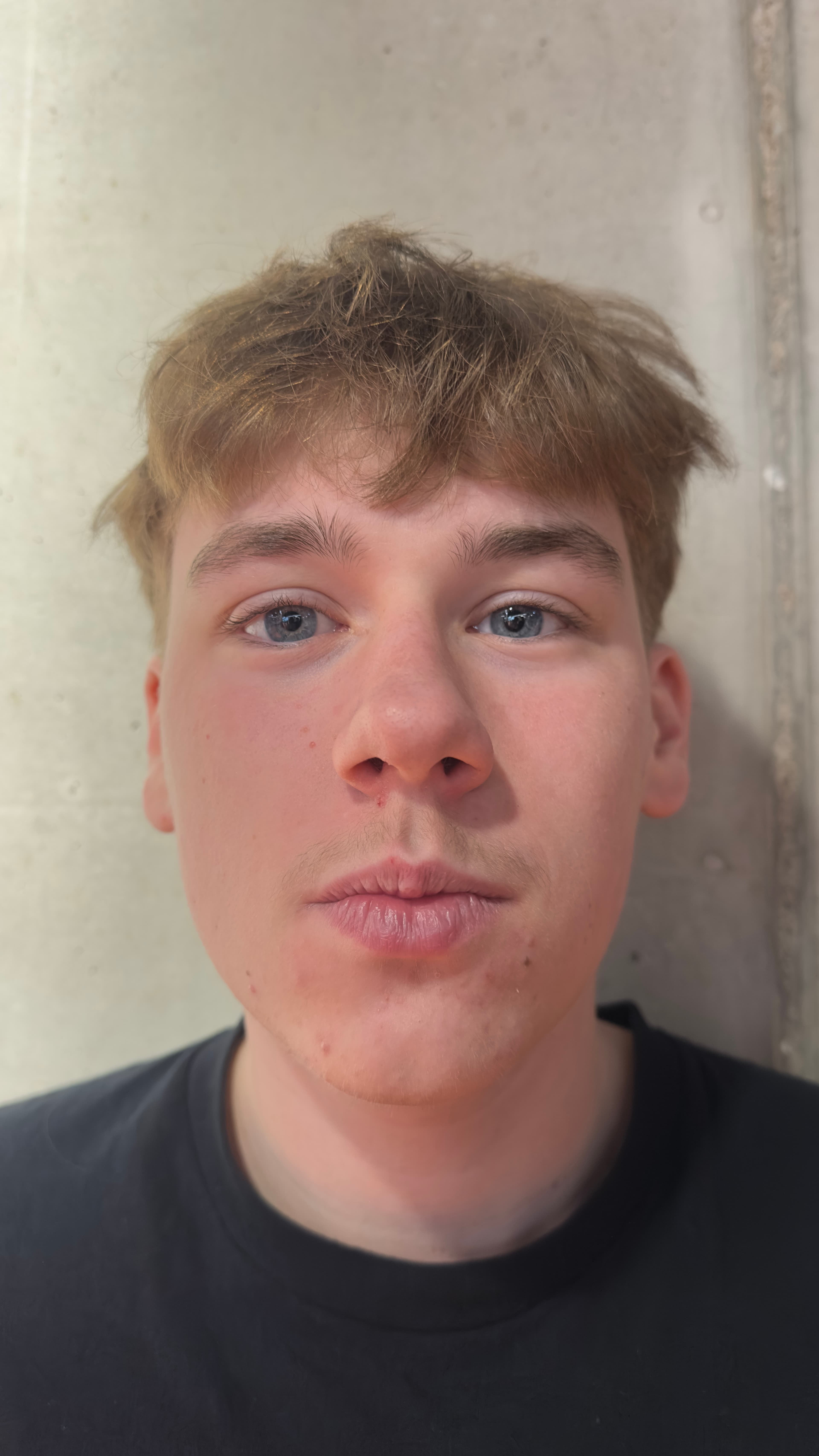 Young man with messy blond hair and blue eyes looking forward against a concrete wall.
