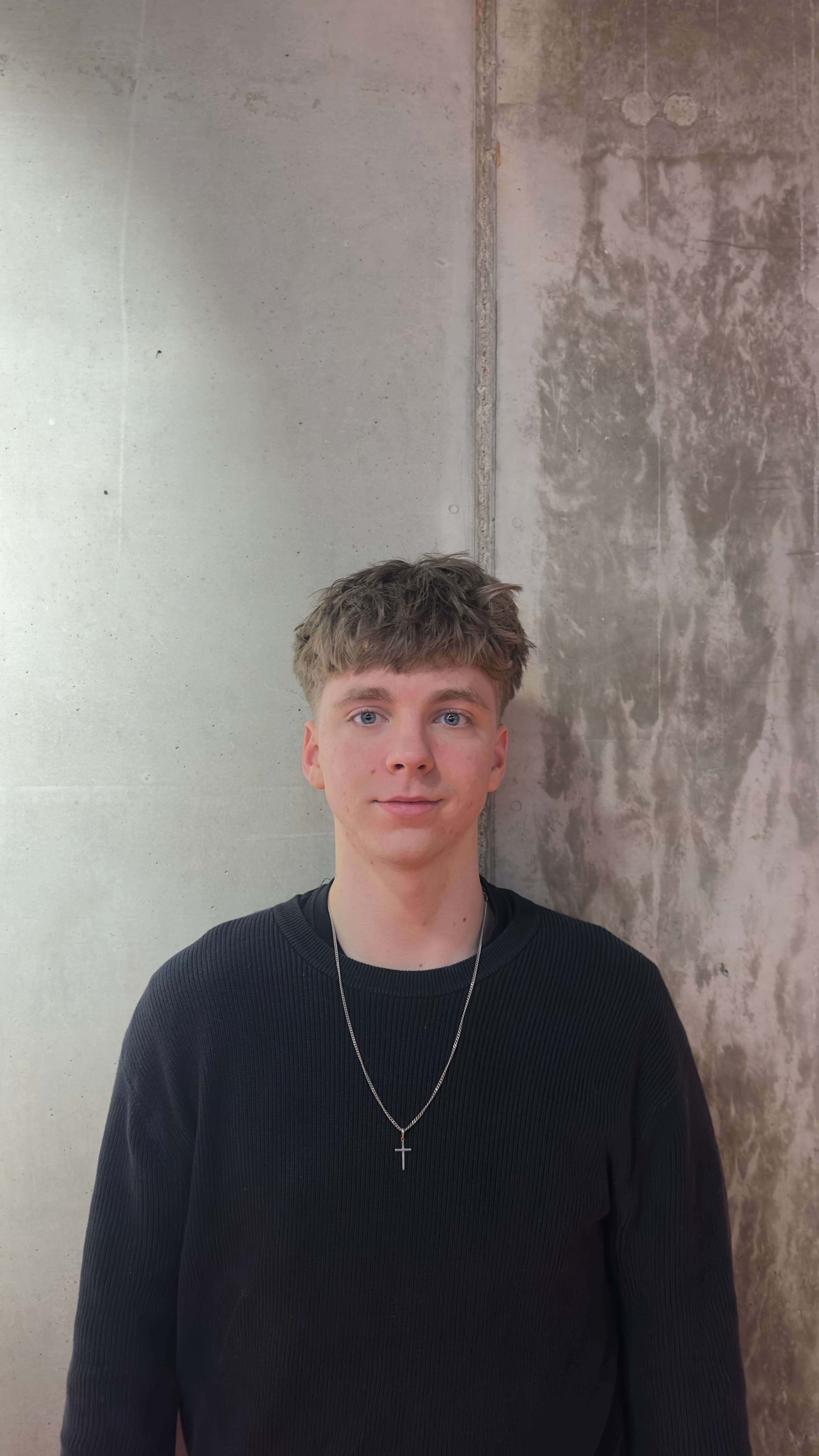 Young man with messy hair and blue eyes wearing a black sweater and cross necklace.